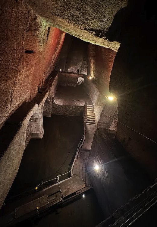 A large Naples Underground cistern lit from above, with dark standing water, walkways and a flight of steps cut into the rock